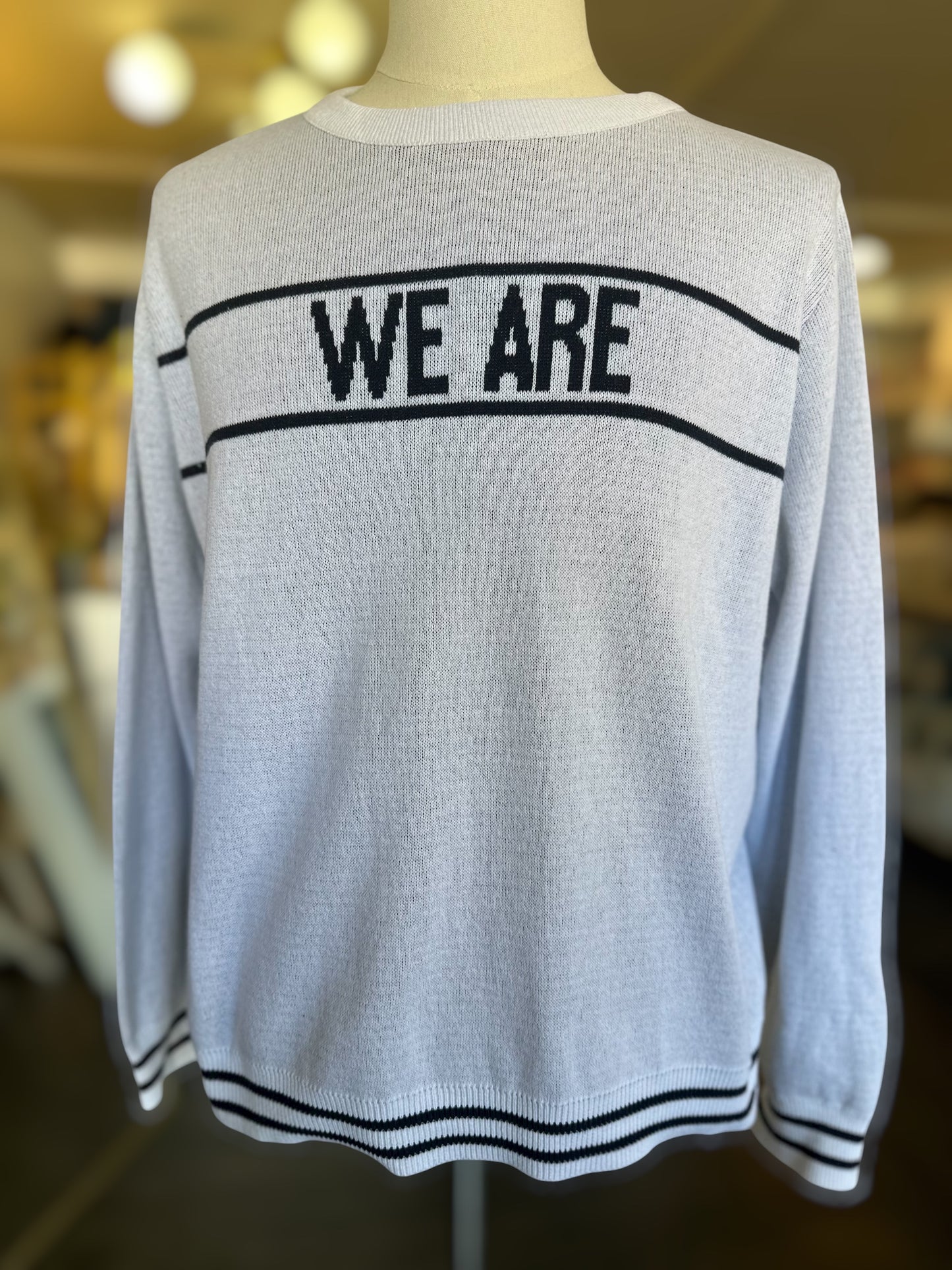 Front-facing lightweight white knit sweater with “WE ARE” text and striped ribbed detailing at the hem and cuffs, shown on a mannequin in a boutique setting