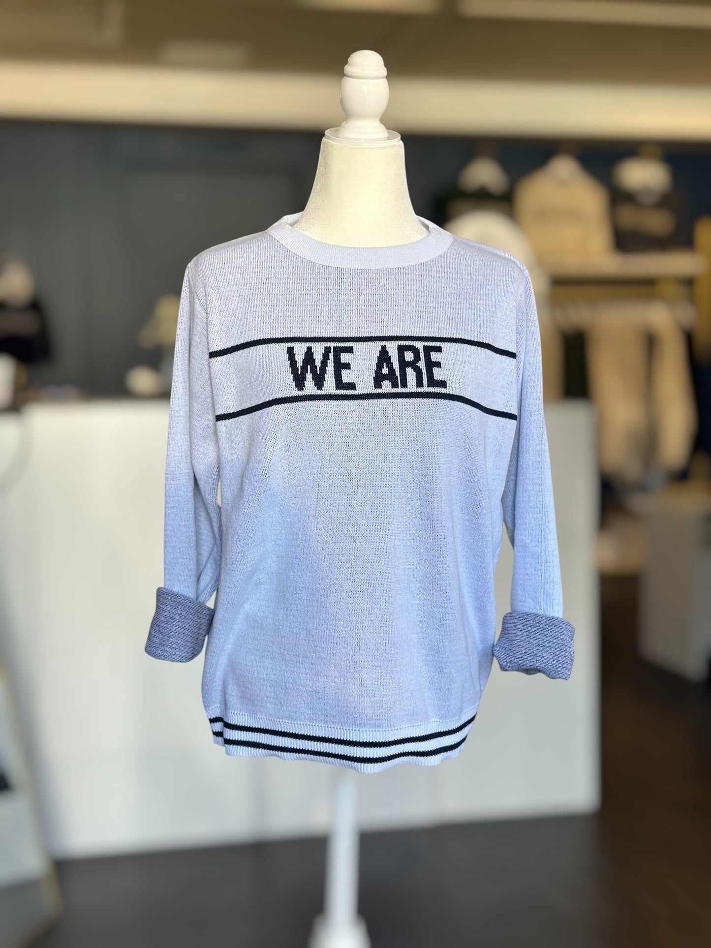 Front view of a lightweight white knit sweater with bold “WE ARE” text across the chest and rolled-up sleeves, displayed on a mannequin.