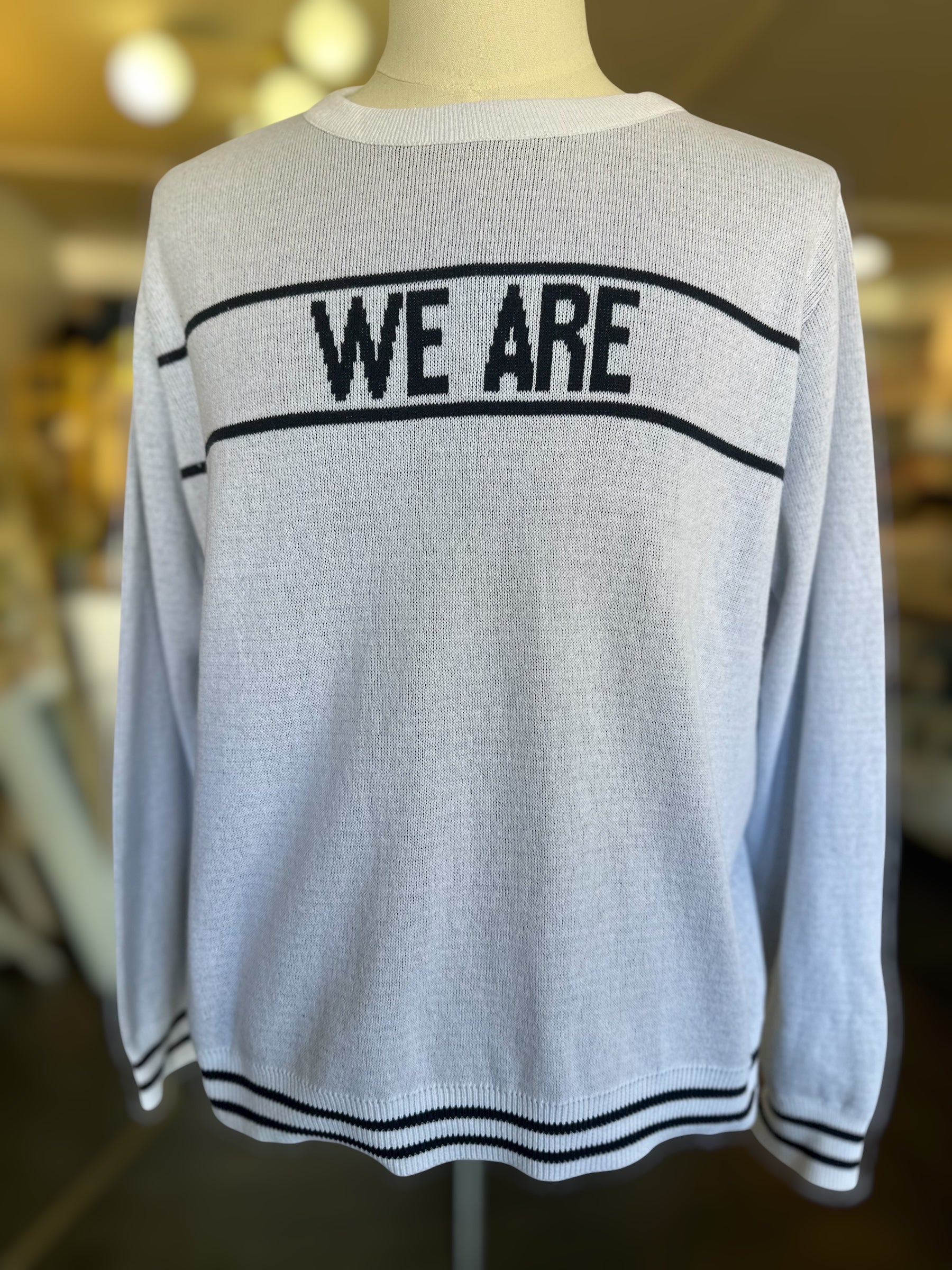 Front-facing lightweight white knit sweater with “WE ARE” text and striped ribbed detailing at the hem and cuffs, shown on a mannequin in a boutique setting